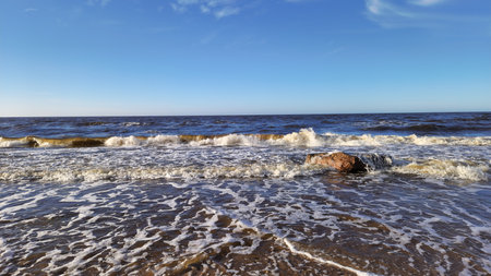 Dynamic view of waves crashing over a solitary rock on a sandy beach under a clear blue sky.の写真素材