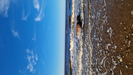 Waves washing over a rocky outcrop on a sunny beach, captured under a blue sky with light clouds.の写真素材