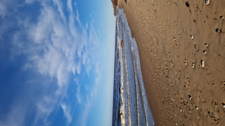 Wide-angle view of a beach scattered with pebbles, leading to the sea under a vast blue sky with wispy clouds.の写真素材