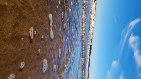 Sunlight dances on foamy waves as they rush towards a sandy beach, framed by a strikingly blue sky.の写真素材