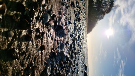 Sunset view of a rocky beach with glistening stones and sparkling water under a bright sun and clear sky.の写真素材