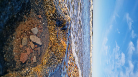 A large, textured rock on the beach foreground with gentle waves and a clear blue sky in the background.の写真素材