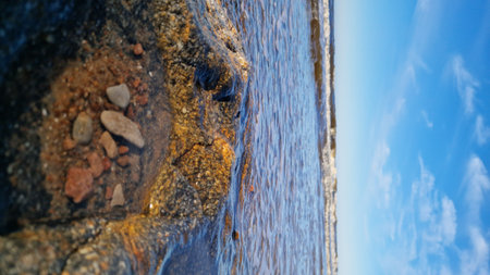 Soft evening light bathes a large rock and surrounding pebbles on the beach, with gentle waves lapping nearby.の写真素材