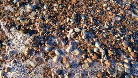 Ocean foam washes over a diverse array of rocks and pebbles at the seaside, captured at sunset.の写真素材