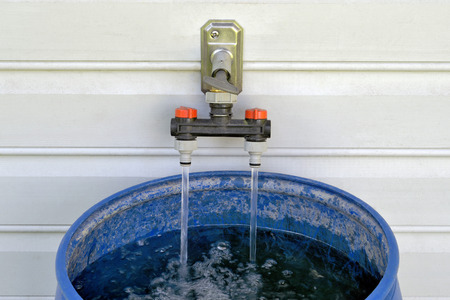 Blue plastic water barrel filling up from water tap on house wall. Purpose: watering the nearby bedding plants using a watering can.の写真素材