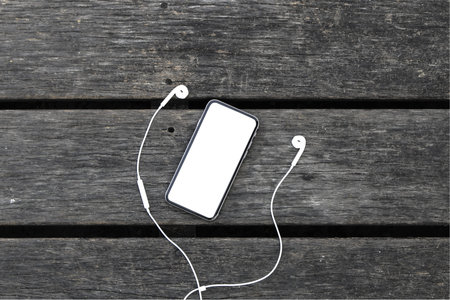 Top view of black mobile smart phone white screen with earphones on wood table background