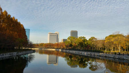 The canal around the Osaka Castle Park. Facing the city area and business buildings. Cloudy blue sky and canal reflection of buildings. Beautiful evening golden sunset light. Osaka, Japan.のeditorial素材