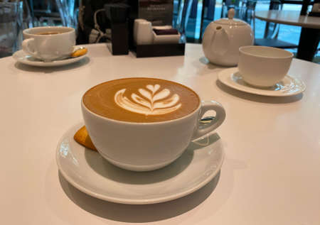 A cup of hot latte with beatiful flower latte art and a piece of cookie. A tea pot in the backgroud.の写真素材