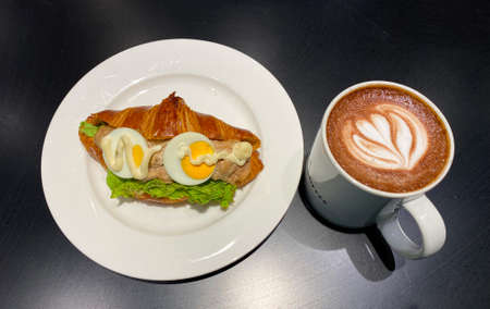 A Cup of fresh hot coffee decorated with flower latte art. With buttery croissant tuna sandwich, filled with savoury mayonnaise tuna, a piece of fresh lettuce and two slices of hard boiled egg.の写真素材
