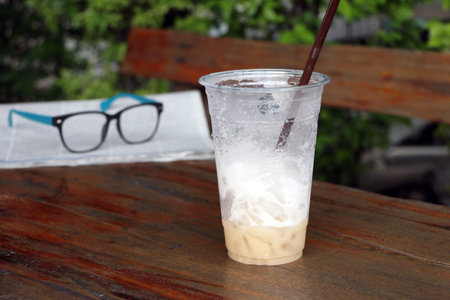 Less cappuccino coffee in the plastic cup and out focus spectacles on the newspaper on the wooden...