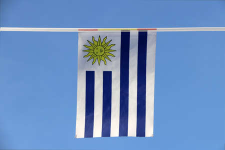Mini fabric rail flag of Uruguay, It has a field of nine equal horizontal stripes alternating white and blue with symbolic of sun. It hanging on the rope cloth on blue sky background.の写真素材
