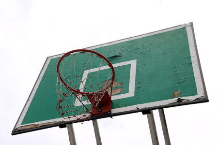 Old green board of basketball with red hoop and white-red mesh on the white sky background....