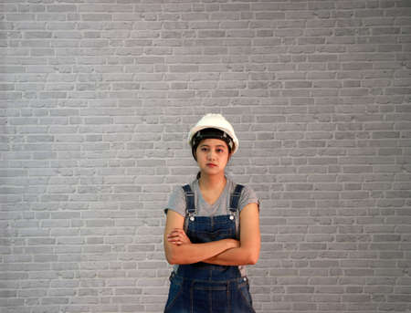 Technician woman ware white helmet with grey T-shirt and denim jeans apron dress standing straight angle and hugging chest on grey brick pattern background.の写真素材