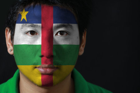 Portrait of a man with the flag of the Central African painted on his face on black background, blue...