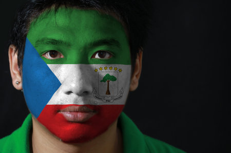 Portrait of a man with the flag of the Equatorial Guinea painted on his face on black background, tricolor of green white and red with a blue triangle and the Coat of arms.の写真素材