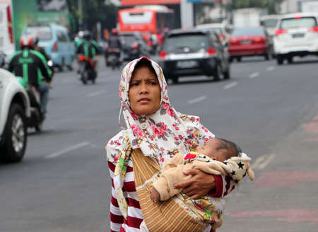 Jakarta, Indonesia May 6, 2019: Muslim woman Indonesian carry children and walking on the footpath beside the road at old town neighborhood in Jakarta.のeditorial素材