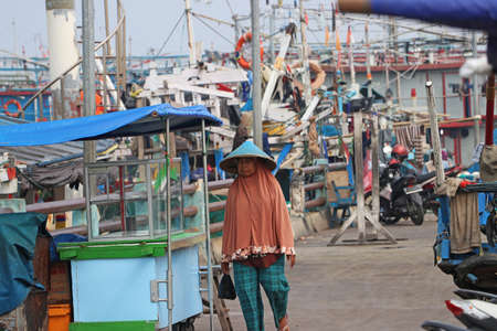 Jakarta, Indonesia May 6, 2019: Muslim woman wearing a conical hat walking at the port harbor which has many fishing boats moored in Jakarta.のeditorial素材