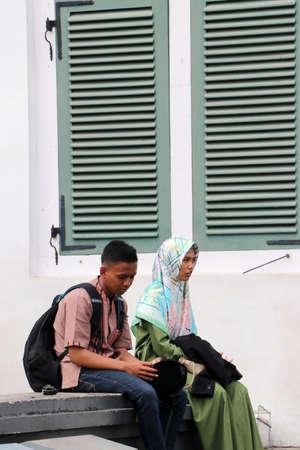 Jakarta, Indonesia May 6, 2019: Muslim male and female teenagers sitting together beside the green window at Fatahillah Square in Old Town.のeditorial素材