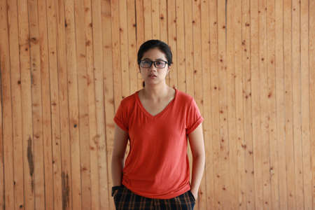Woman wearing orange color of shirt and standing with two hands in pant pockets on the wooden wall background.の写真素材