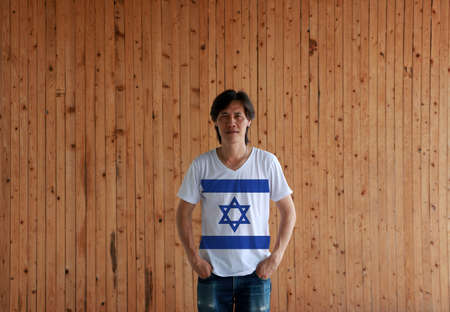 Man wearing Israel flag color shirt and standing with two hands in pant pockets on the wooden wall background, blue hexagram on a white background, between two blue stripes.の写真素材