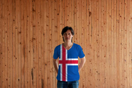 Man wearing Iceland flag color of shirt and standing with crossed behind the back hands on the wooden wall background, blue sky with a snow-white cross and a fiery-red cross inside the white.の写真素材