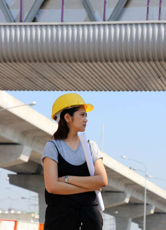 Female civil engineer or architect with yellow helmet, standing and hugging chest with project drafts while in the work area.の写真素材