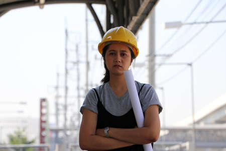 Female civil engineer or architect with yellow helmet, standing and hugging chest with project drafts while in the work area.の写真素材