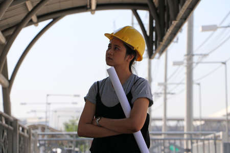 Female civil engineer or architect with yellow helmet, standing and hugging chest with project drafts while in the work area.の写真素材