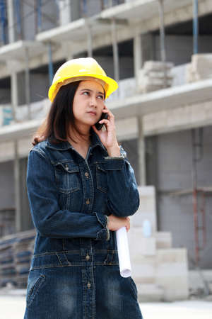 Female civil engineer or architect with yellow helmet, standing and calling with mobile phone and project drafts while in hand at the work area.の写真素材