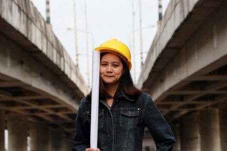 Female civil engineer or architect with yellow helmet, standing with project drafts while in hand on parallel expressway background.の写真素材