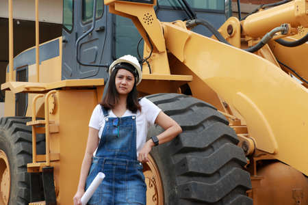 Female civil engineer or architect with white helmet and project drafts while in hand standing with Alloy Bulldozer Truck with Front Loader Truck Engineering Construction Car Vehicle at the work area.の写真素材