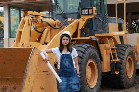 Female civil engineer or architect with white helmet and project drafts while in hand standing with Alloy Bulldozer Truck with Front Loader Truck Engineering Construction Car Vehicle at the work area.の写真素材