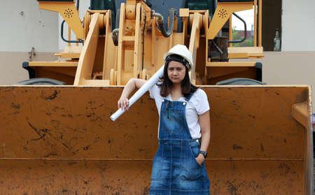 Female civil engineer or architect with white helmet and project drafts while in hand standing with Alloy Bulldozer Truck with Front Loader Truck Engineering Construction Car Vehicle at the work area.の写真素材