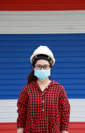 Female civil engineer or architect with white helmet and masked prevent germs, wear red plaid dress on the color of Thailand flag door background. Tiny Particle or virus corona or Covid 19 protection.の写真素材