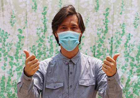 Masked Asian man prevent germs and wear blue long sleeved shirt with thumbs up. Tiny Particle or...