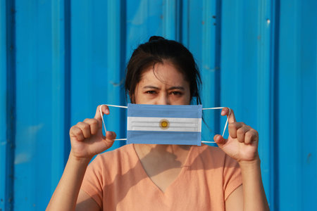 A woman with Argentina flag on hygienic mask in her hand and lifted up the front face on blue background. Tiny Particle or virus corona or Covid 19 protection. Concept of Combating illness.の写真素材