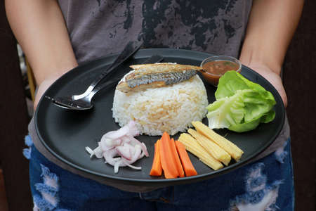 Hand served breakfast in the black round plate. Fried mackerel and Rice with Spicy Shrimp Paste Dip and vegetable, Chinese cabbage, baby corn, carrots and sliced onions with spoon and fork.  の写真素材