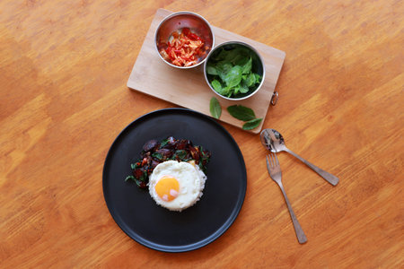 Chinese sausage fried with basil and fried egg with rice in the black round dish, Cooking ingredients with spoon and fork on the wooden floor.の写真素材