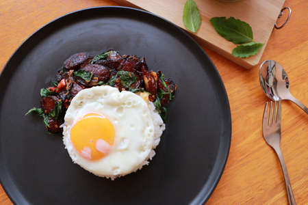 Chinese sausage fried with basil and fried egg with rice in the black round dish, Cooking ingredients with spoon and fork on the wooden floor.の写真素材