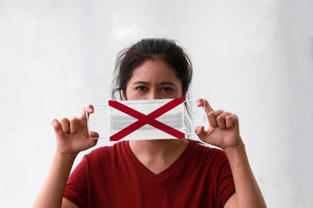 A woman and hygienic mask with Alabama flag pattern in her hand and raises it to cover her face. A mask is a very good protection from Tiny Particle or virus corona or Covid 19.の写真素材