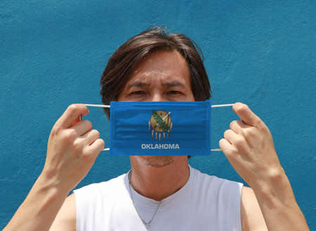 A man and hygienic mask with Oklahoma flag pattern in his hand and raises it to cover his face. A mask is a very good protection from Tiny Particle or virus corona or Covid 19.の写真素材
