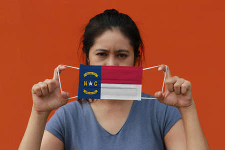 A woman and hygienic mask with North Carolina flag pattern in her hand and raises it to cover her face on orange color background. A mask is a very good protection from Tiny Particle or virus corona or Covid 19.の写真素材