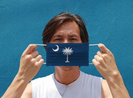 A man and hygienic mask with South Carolina flag pattern in his hand and raises it to cover his face on blue background. A mask is a very good protection from Tiny Particle or virus corona or Covid 19.の写真素材