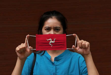 A woman and hygienic mask with Isle of man flag pattern in her hand and raises it to cover her face on dark background. A mask is a very good protection from Tiny Particle or virus corona or Covid 19.の写真素材