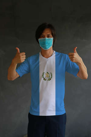 Masked man wearing Guatemala flag color of shirt and thumbs up with both hands on dark wall background. Concept of protection and fighting COVID.の写真素材