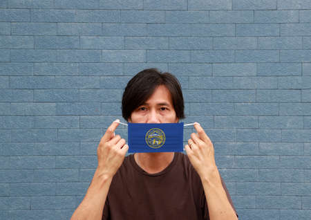 A man and hygienic mask with Nebraska flag pattern in his hand and raises it to cover his face. A mask is a very good protection from Tiny Particle or virus corona or Covid 19.の写真素材