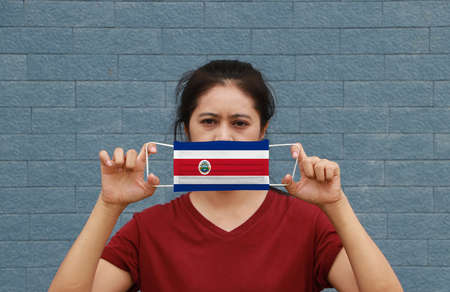 A woman and hygienic mask with Costa Rica flag pattern in her hand and raises it to cover her face on blue background. A mask is a very good protection from Tiny Particle or virus corona or Covid 19.の写真素材
