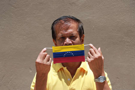 A man with Venezuela flag on hygienic mask in his hand and lifted up the front face and he wear yellow shirt. Tiny Particle or virus corona or Covid 19 protection. Concept of Combating illness.の写真素材