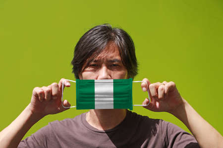 A man and hygienic mask with Nigeria flag pattern in his hand and raises it to cover his face on green background. A mask is a very good protection from Tiny Particle or virus corona or Covid 19.の写真素材