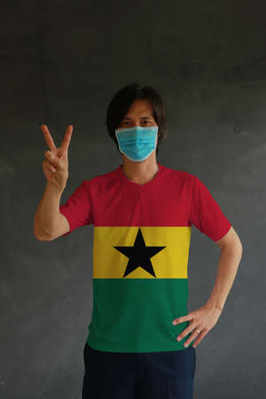 Man wearing hygienic mask and wearing Ghanaian flag colored shirt and raising two fingers up on dark wall background. Concept of protect tiny dust or disease of Ghana.の写真素材